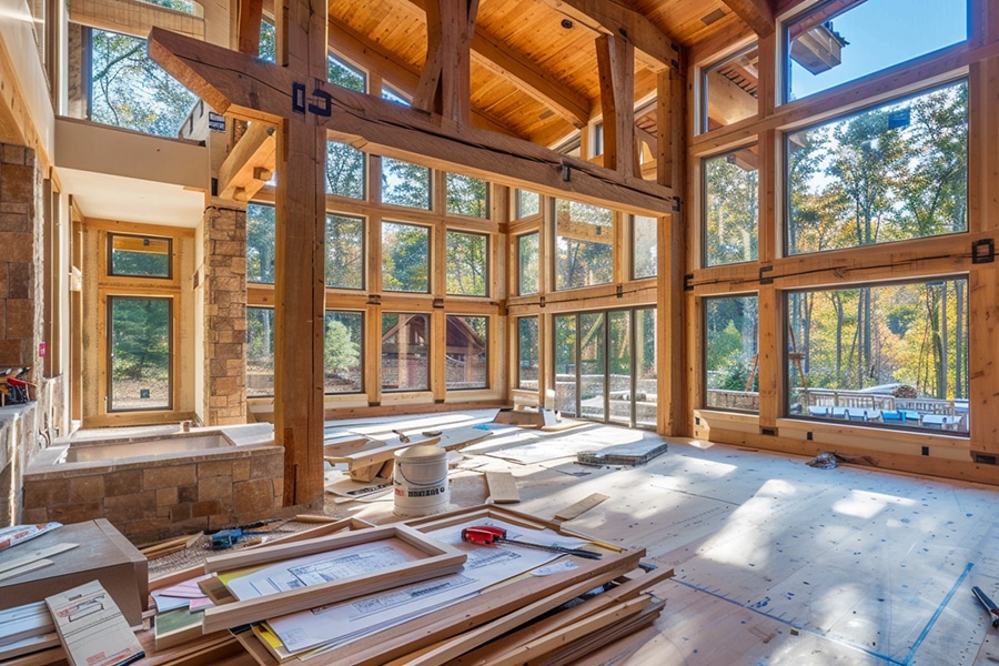 Large, unfinished wooden interior with high ceilings, exposed beams, and tall windows overlooking a wooded area; construction materials and blueprints scattered on the floor.